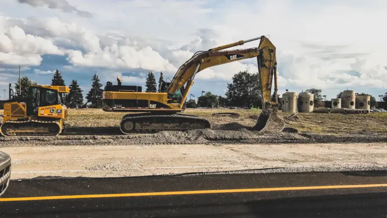 Ein gelber Bagger bei Bauarbeiten neben einer befahrenen Straße bei sonnigem Wetter.