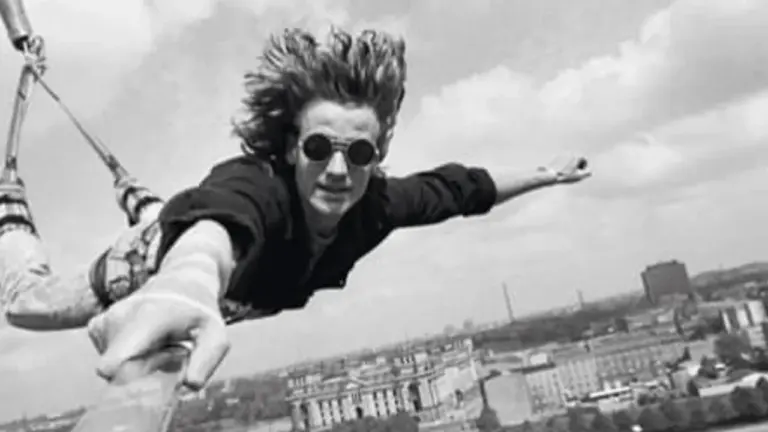 Ein junger Mann mit Sonnenbrille springt kopfüber beim Bungee-Jumping vor einer urbanen Kulisse in die Tiefe.