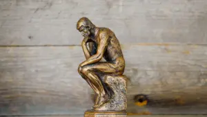 Eine kleine bronzene Denker-Statue steht nachdenklich vor einem rustikalen Hintergrund aus Holz.