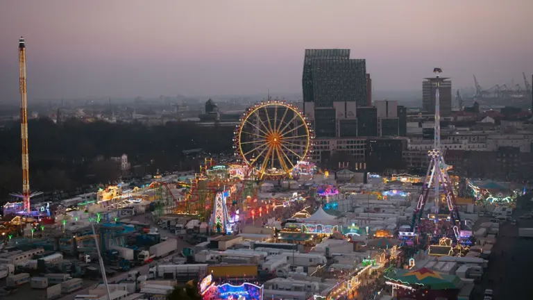 Weitwinkelaufnahme des Hamburger Doms bei Abenddämmerung mit beleuchtetem Riesenrad und Skyline.