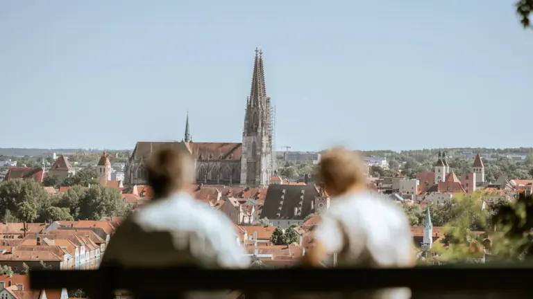 Zwei Personen blicken von einer Bank auf den Regensburger Dom und die Altstadt.