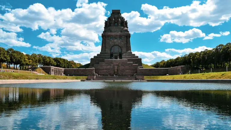 Das massive Völkerschlachtdenkmal in Leipzig spiegelt sich bei blauem Himmel im Wasserbecken.